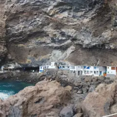 Casas blancas con detalles azules se asoman desde una cueva en los poris de Candelaria, La Palma. El mar turquesa rompe contra las rocas.
