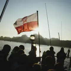 Słońce przebija się przez biało-czerwoną bandera polską na statku. Grupa ludzi obserwuje zachód słońca nad wodą.