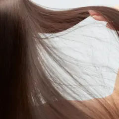 Mano acariciando cabello castaño, fino y escaso, que cae en cascada sobre un fondo blanco.
