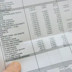 Une main tient une fiche de paie perdue, détaillant les salaires et les retenues.