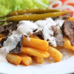 En tallrik med pommes frites, kebabkött och vit sås. Tillbehör som sallad, gurka, tomat, lök och jalapeños. Ett gott kebab recept!