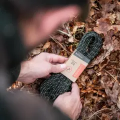 Manos sostienen un rollo de cordón paracord verde con puntos blancos, sobre un lecho de hojas secas.