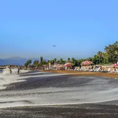 Ein sonniger Antalya Strand mit sanften Wellen, Liegestühlen und Palmen. Ein Flugzeug zieht seine Bahn am blauen Himmel.