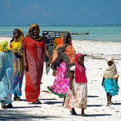 Grupa kobiet i dziewcząt w kolorowych strojach spaceruje po piaszczystej plaży z widokiem na ocean.