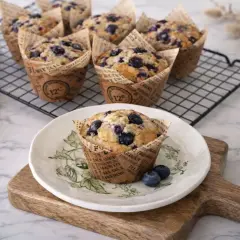 Un délicieux muffin myrtille, tout juste sorti du four, posé sur une assiette décorée. D'autres muffins myrtilles refroidissent sur une grille.