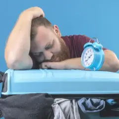 Zmęczony podróżnik śpi na walizce, budzik obok sygnalizuje jetlag.