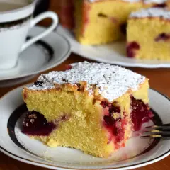 Szybkie ciasto bezglutenowe z wiśniami, posypane cukrem pudrem, na talerzyku obok filiżanki kawy. Idealne na deser.