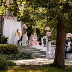 Piękny ślub kościelny w plenerze. Para młoda siedzi przed ołtarzem, a ksiądz udziela im błogosławieństwa. Wokół kwitną kwiaty i drzewa.