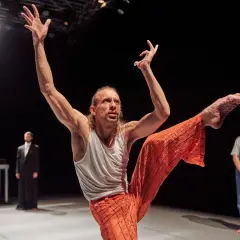 Tancerz w pomarańczowych spodniach wykonuje dynamiczny ruch, prezentując sztukę na wynos. W tle widzimy innych performerów.