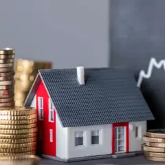 Casa modelo rodeada de pilas de monedas, con un gráfico ascendente al fondo, sugiriendo un estudio de solvencia y el valor de la inversión inmobiliaria.
