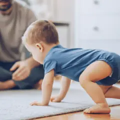 Dziecko w niebieskim body raczkuje po dywanie, obok siedzi ojciec z zabawkami.