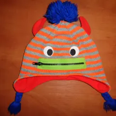 Pomarańczowo-szara czapka z rogami, niebieskim pomponem i zielonym zamkiem imitującym usta.
