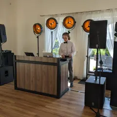 Młody DJ w muszce i białej koszuli obsługuje sprzęt muzyczny przy drewnianym stanowisku, otoczony głośnikami i oświetleniem.