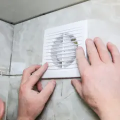 Installation d'un ventilateur pour l'aération de la salle de bain. Des mains ajustent l'appareil blanc sur un mur carrelé.