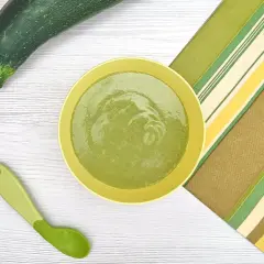 Un plato con puré de calabacín bebé, una cuchara verde y un calabacín entero sobre una mesa de madera.