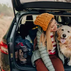 Uśmiechnięta dziewczyna w żółtej czapce przytula swojego złotego retrievera w bagażniku auta. W tle buty trekkingowe i ubrania, sugerujące wycieczkę. To idealny przykład habituacji psa do podróży.