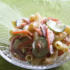 A close-up of a crystal bowl filled with creamy Japanese macaroni salad, featuring elbow macaroni, shredded carrots, sliced cucumbers, and ham.