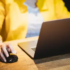 Kobieta w żółtej marynarce klika myszką przy laptopie, porządkując sprawy, być może rachunki za prąd po śmierci bliskiej osoby.