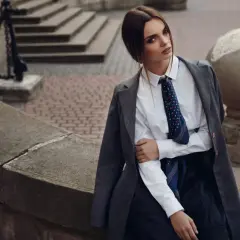Elegancka kobieta w białej bluzce z krawatką i szarej marynarce, siedząca na kamiennej ławce.