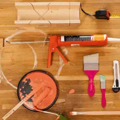 Un tableau peinture acrylique sur bois, prêt pour un projet de bricolage. Pinceaux, rouleau, pot de peinture orange, pistolet à mastic, ruban adhésif, mètre et équerre sont disposés sur un plan de travail en bois.