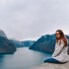 Kobieta siedzi na skarpie z widokiem na norweski fiord i ośnieżone góry.