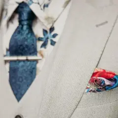 Elegancki mężczyzna w jasnej marynarce, z niebieskim wzorzystym krawatem i spinką do krawata jak nosić. W kieszeni kolorowa poszetka.