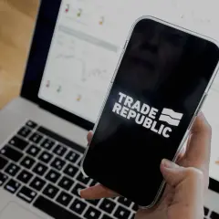 Person prüft den Trade Republic Quartalsbericht auf dem Smartphone, während der Laptop mit Finanzdiagrammen im Hintergrund läuft.
