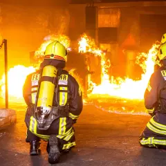 Zwei Feuerwehrleute in voller Montur knien vor einem intensiven Feuer. Ihre Ausbildung bei der Berufsfeuerwehr bereitet sie auf solche Einsätze vor.
