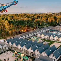 Widok z lotu ptaka na holiday park z domkami, placem zabaw i basenem, a nad nim latający helikopter.