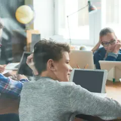 Kinder lernen mit Tablets und Laptops in einem modernen, hellen Raum. Eine Tafel mit mathematischen Formeln deutet auf ein digitales Klassenzimmer mit App-gestütztem Unterricht hin.