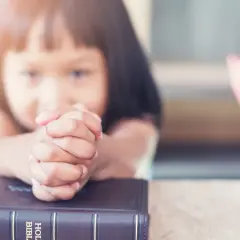 Dziecko z dłońmi złożonymi do modlitwy nad Biblią. Obok kolorowe kredki, symbol nauki dekalogu 10 przykazań.