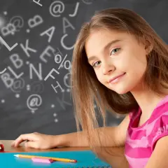 Een meisje met een roze gestreepte trui zit aan een bureau, omringd door zwevende letters en symbolen, klaar om woordjes te leren.