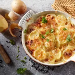 Ziemniaki po francusku, zapiekane w kremowym sosie, posypane natką pietruszki. Obok surowe ziemniaki i nóż.