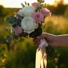 Dłoń panny młodej trzyma bukiet róż i eukaliptusa, ozdobiony satynową wstążką. Różne rodzaje wstążek florystycznych dodają uroku.