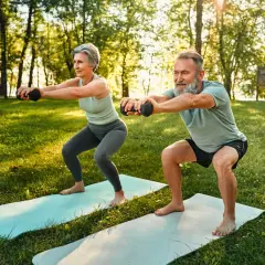 Une femme sportive de 50 ans et un homme font des squats avec des haltères dans un parc ensoleillé.