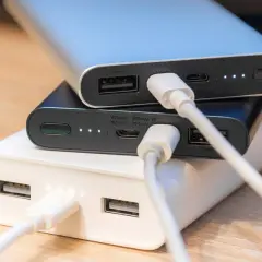 Trzy powerbanki w kolorach białym, czarnym i srebrnym, podłączone kablami USB. To przenośne ładowarki, które pomogą naładować urządzenia w podróży.
