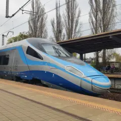 Nowoczesny eip pociąg Pendolino stoi na peronie. W tle widać wiatę, drzewa i budynki.