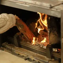 Main tenant ajoutant une bûche dans un poêle à bois, les flammes dansent joyeusement.