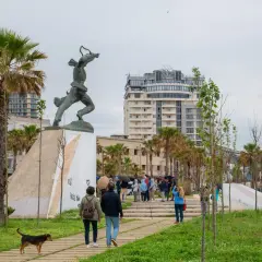 Pomnik i ludzie spacerujący po parku w Durrës. W tle nowoczesne budynki i palmy.