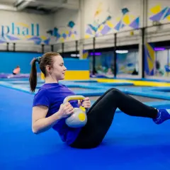 Kobieta ćwiczy z kettlebell na niebieskiej macie w sali gimnastycznej z trampolinami w tle.