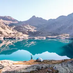 Un randonneur admire un lac turquoise aux reflets parfaits dans le **parc du Mercantour**. Une tente est installée sur la rive rocheuse.