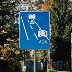 Niebieski znak drogowy z białymi symbolami aparatów fotograficznych i falami, informujący o odcinkowym pomiarze prędkości.