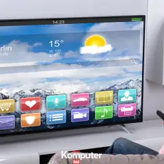 Kobieta z pilotem ogląda nowoczesny telewizor LCD, na którym wyświetla się interfejs z ikonami aplikacji i prognozą pogody dla Berlina.
