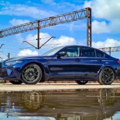 Granatowe BMW M3 G80 stoi obok torów kolejowych, odbijając się w kałuży.
