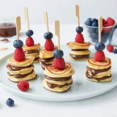 Mini pancakes rezept: gestapelte Mini-Pfannkuchen mit Schokocreme, garniert mit Himbeeren und Blaubeeren auf Spießen.