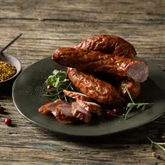 Na talerzu ułożona jest pyszna, wędzona kiełbasa z dziczyzny, pokrojona w plastry. Obok miseczka z musztardą ziarnistą, żurawina i gałązki rozmarynu.