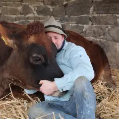 Ein Mann in Jeans und kariertem Hemd umarmt innig eine braune Kuh. Sie kuscheln im Stroh vor einer Steinmauer.