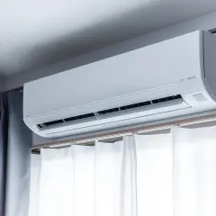 Biały klimatyzator nad oknem, z zasłonami po bokach, zapewnia chłód w upalne dni.