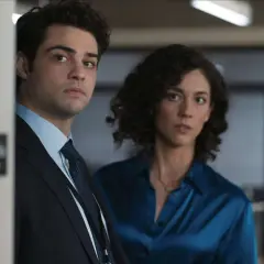 Besetzung von The Recruit: Ein Mann im Anzug und eine Frau in einem blauen Hemd blicken ernst in die Kamera.