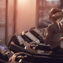 Frau mit blondem Pferdeschwanz walkt auf dem Laufband im Fitnessstudio, das warme Abendlicht fällt durch die Fenster.